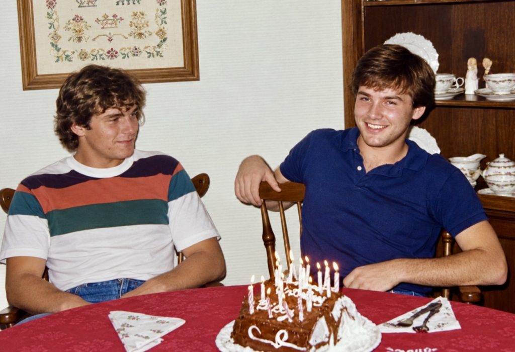 Two young men seated at a dining table behind a chocolate birthday cake with lit candles. One, in a striped T-shirt, looks toward the other, who wears a navy polo shirt and smiles at the camera.